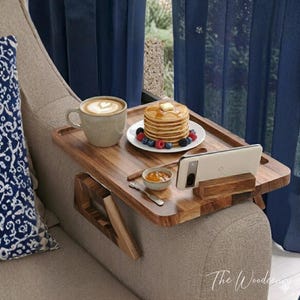 Könnte beinhalten: Ein Holz-Sofa-Arm-Tablett mit einer Tasse Kaffee, Pfannkuchen mit Beeren, einer kleinen Schale Marmelade und einem Smartphone. Das Tablett aus dunklem Holz hat einen eingebauten Telefonständer. Das Tablett liegt auf einem beigen Sofa.