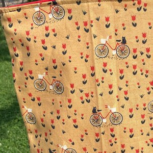 Peut inclure: Un sac fourre-tout beige orné d'un motif répété de vélos rouges avec des chats noirs et de tulipes rouges. Il possède des anses noires et une bordure rouge.
