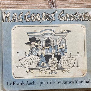 May include: A blue book cover with the title "Mac Goose's Grocery" in white lettering. The cover features a cartoon illustration of a family of geese in front of a store with a sign that reads "Mac Goose's". The book is by Frank Asch with pictures by James Marshall.