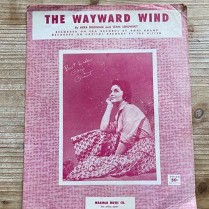 May include: A pink sheet music cover for the song "The Wayward Wind" by Herb Newman and Stan Lebowsky. The cover features a photo of a woman sitting on the ground, looking off to the side. The text "Best wishes Gogi Grant" is handwritten in black ink on the cover.