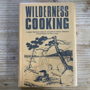 May include: A tan-colored book with the title "WILDERNESS COOKING" in black lettering. The book cover features a black and white illustration of a campfire with smoke rising from it, surrounded by trees and a lake.