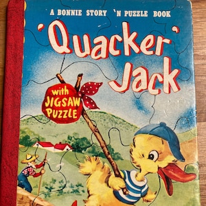 May include: A vintage children's book titled "'Quacker' Jack" with a jigsaw puzzle. The cover features a cartoon duck wearing a blue and white striped shirt and a blue cap, holding a stick with a red bow and a red apple with the words "with jigsaw puzzle" on it. The duck is running on a green hill with a red roof house in the background.