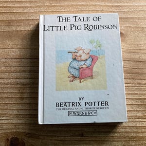 May include: A hardcover book titled The Tale of Little Pig Robinson by Beatrix Potter. The white cover features a central illustration of a pig sitting in a red chair on a beach, looking through a telescope. The text includes the author's name and F. Warne & Co.