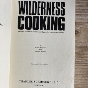 May include: A vintage book cover with the title "WILDERNESS COOKING" in bold black text. The book is a unique illustrated cookbook and guide for outdoor enthusiasts by Berndt Berglund and Clare E. Bolsby. It is illustrated by E.B. Sanders and published by Charles Scribner's Sons in New York.