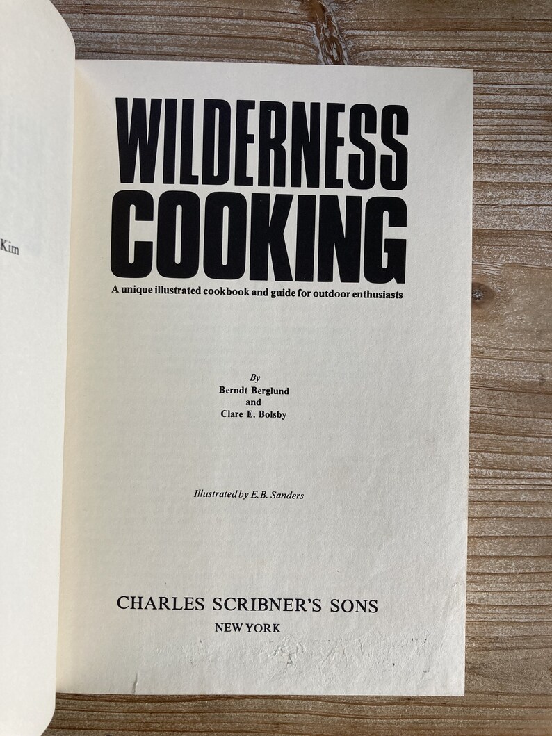 May include: A vintage book cover with the title "WILDERNESS COOKING" in bold black text. The book is a unique illustrated cookbook and guide for outdoor enthusiasts by Berndt Berglund and Clare E. Bolsby. It is illustrated by E.B. Sanders and published by Charles Scribner's Sons in New York.