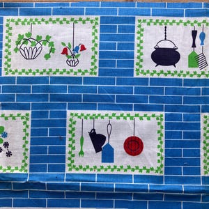 Op de afbeelding: Een blauw en wit keukenstof met een baksteenpatroon. De stof heeft rechthoekige panelen met illustraties van keukenartikelen zoals hangplanten, een theepot en een ketel. De illustraties zijn omlijst met groene wijnranken.