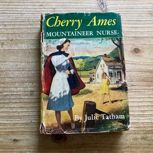 May include: Vintage book cover for "Cherry Ames, Mountaineer Nurse" by Julie Tatham. The cover features an illustration of a nurse in a white uniform and red cape, with a young girl in a yellow dress. The background shows a house and a rural landscape.