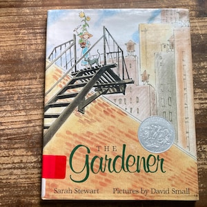 May include: A hardcover book titled The Gardener by Sarah Stewart with pictures by David Small. The cover features an illustration of a person with a potted plant and a cat on a fire escape against a city skyline, with a silver award seal.
