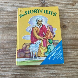May include: A yellow book titled "The Story of Jesus" with a blue and white border. The cover features an illustration of Jesus with two children and a lamb. The book is a first religious book edited by Dr. Mary Alice Jones.