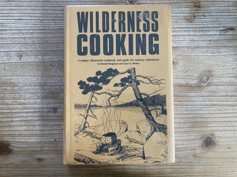 May include: A tan-colored book with the title "WILDERNESS COOKING" in black lettering. The book cover features a black and white illustration of a campfire with smoke rising from it, surrounded by trees and a lake.