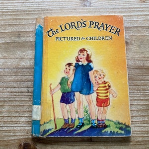 May include: A vintage children's book titled "The Lord's Prayer Pictured for Children". The cover features a yellow background with three children, two boys and a girl, standing together. The girl is wearing a blue dress and the boys are wearing green and yellow striped shirts.