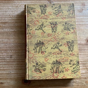 May include: A hardcover book with a tan cover featuring a repeating pattern of black line-art illustrations of figures, birds, and stars, accented by thin red sunburst designs. The book has a dark brown spine and a textured, wood-grain appearance.