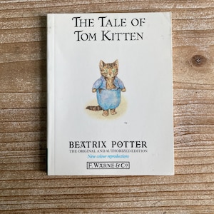 Peut inclure: Une couverture de livre vintage avec une illustration de dessin animé d'un chaton brun et blanc portant une chemise bleue. Le titre du livre est "The Tale of Tom Kitten" de Beatrix Potter. La couverture du livre est blanche avec du texte noir.