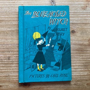 May include: A hardcover book titled The Bluenosed Witch by Margaret Embry with pictures by Carl Rose. The teal cover features a black and white illustration of a girl in a dress and yellow boots holding a witch hat, a black cat, and a yellow moon.