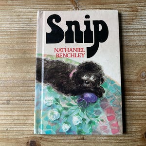 May include: A book titled "Snip" by Nathaniel Benchley. The cover features a black dog with a purple flower in its mouth, lying on a colorful floral patterned background.