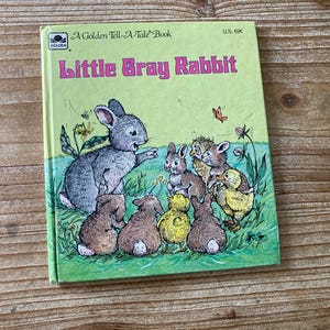 May include: A Golden Tell-A-Tale Book titled "Little Gray Rabbit" features a gray rabbit and other animals. The cover has a light green background with illustrations of the characters. The book is on a wooden surface.