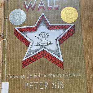 May include: A hardcover book titled The Wall: Growing Up Behind the Iron Curtain by Peter Sís. The cover features a red brick star illustration with a child playing a drum inside. It includes a silver Caldecott Honor seal and a gold Sibert Medal.