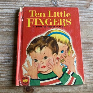 May include: A red hardcover book titled "Ten Little Fingers: A Book of Finger Plays" with illustrations of two children. The boy is wearing a green and red striped shirt and the girl is wearing a blue headband and a white dress.