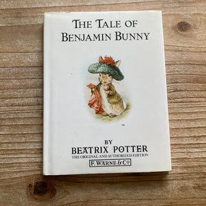 May include: A hardcover book titled "The Tale of Benjamin Bunny" by Beatrix Potter. The cover features an illustration of a rabbit wearing a hat and holding a red cloth. The book is on a wooden surface.