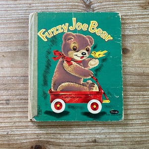 May include: Vintage children's book titled "Fuzzy Joe Bear." The cover features a brown bear with a red bow, sitting in a red wagon, and holding a yellow bird. The background is teal with the title in yellow.