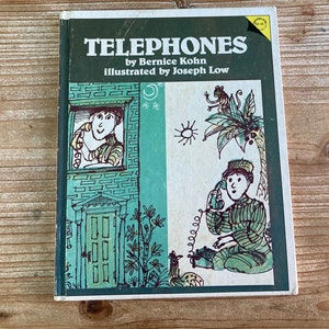 May include: A vintage children's book titled "Telephones" by Bernice Kohn, illustrated by Joseph Low. The cover features illustrations of people using telephones, with a green and white color scheme. The book is set on a wooden surface.