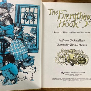 May include: Open book with illustrations and text. The left page features a blue-toned illustration of a family scene with children and a cat. The right page displays the title "The Everything Book" and author information.