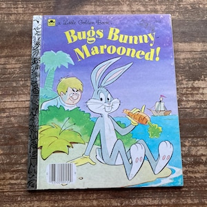 May include: A Little Golden Book cover featuring Bugs Bunny, a cartoon rabbit, holding a carrot. The book title is "Bugs Bunny Marooned!" and the cover is illustrated in a cartoon style with a purple background and a blue sky.