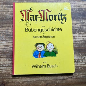 May include: A yellow hardcover book titled Max und Moritz, eine Bubengeschichte in sieben Streichen by Wilhelm Busch. The cover features a black gothic-style title and a colorful illustration of two boys in blue and green shirts on a yellow background.