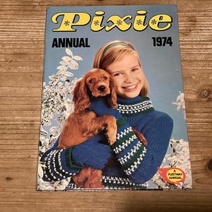 May include: A vintage "Pixie Annual 1974" book cover. The cover features a smiling girl holding a brown dog, both wearing blue sweaters. The title is in yellow with snowflake accents. A Fleetway Annual logo is in the bottom right corner.