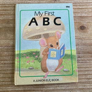 May include: A children's book titled "My First ABC" featuring a cartoon mouse reading a book under a mushroom. The book cover is blue and white with the title "My First ABC" in a blue font. The mouse is brown with a pink nose and is wearing a blue and white book.
