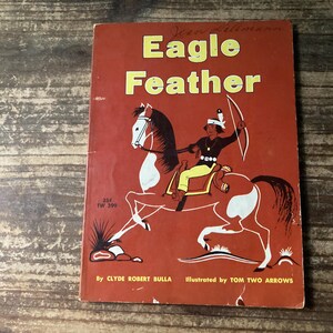 May include: A vintage paperback book titled Eagle Feather by Clyde Robert Bulla, illustrated by Tom Two Arrows. The cover is rust-colored with a graphic illustration of a person riding a pinto horse while holding a bow and arrow.