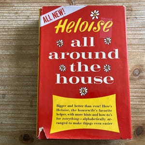 May include: A red book with a yellow title that reads "Heloise all around the house". The book has a white daisy design and the text "All New!" in the top left corner. The book is on a wooden surface.