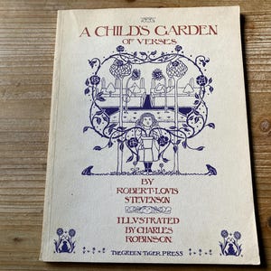 May include: A vintage book titled "A Child's Garden of Verses" by Robert Louis Stevenson. The cover features a detailed illustration in blue and white, with floral and whimsical elements. The book is on a wooden surface.