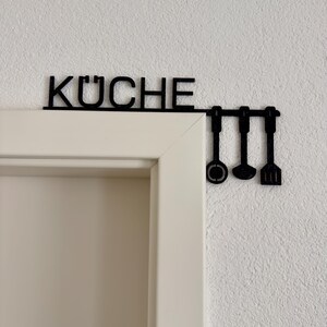 May include: Black metal kitchen sign that reads "KÜCHE" above a door frame, with three utensil-shaped hooks hanging below. The sign is mounted on a white wall.
