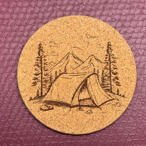 以下が含まれることがあります： 山岳風景の中にテントがレーザー彫刻されたコルクコースター。デザインは、開いたフラップのテント、背景の山々、両側の木々を特徴としています。