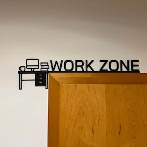 Può includere: Una silhouette in bianco e nero di una scrivania con un computer, una tazza e libri, con il testo "WORK ZONE" sopra. La silhouette è su un muro bianco, e una porta di legno è a destra.