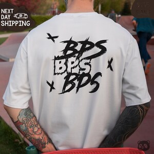 May include: White t-shirt with a black graphic design of the word "BPS" repeated three times in a stylized font. The graphic is surrounded by small black crosses.