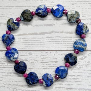 May include: A beaded bracelet featuring faceted heart-shaped lapis lazuli stones in shades of deep blue and grey, interspersed with small, round, faceted magenta-colored beads arranged in a circular pattern on a white wood-grain surface.
