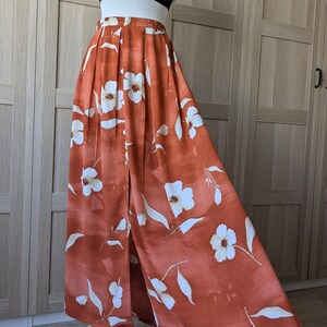Op de afbeelding: Een zwarte crop top met spaghettibandjes gecombineerd met een rok met hoge taille die tot op de grond valt in een roestoranje kleur. De rok heeft een witte bloemenprint, een knoopsluiting aan de voorzijde en verticale plooien bij de tailleband.