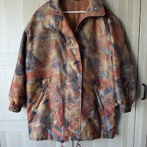 Op de afbeelding: Een vintage, oversized jas met een abstract patroon in bruin, oranje en blauw. De jas heeft een kraag, knoopsluitingen en twee voorzakken. Hij hangt aan een houten hanger.