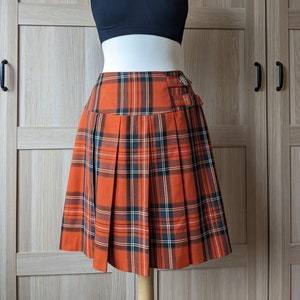 Pode incluir: Um top preto e uma saia plissada xadrez em tons de laranja, preto, branco e azul marinho. A saia apresenta dois fechos de fivela e é exibida em um manequim. A roupa é adequada para uso casual.