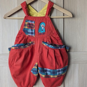 Op de afbeelding: Rode kinderoverall met gele voering en een clownpatch. De overall heeft geruite accenten en een houten hanger. De totale lengte is ongeveer 46 cm.