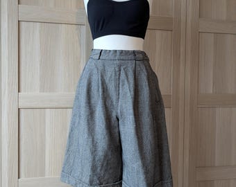 May include: A black sports bra and gray plaid culottes displayed on a mannequin. The culottes have a high waist and wide legs, with a subtle checkered pattern. The outfit is styled on a wooden stand against a light-colored wooden backdrop.