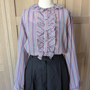 以下が含まれることがあります： マネキンに展示された、フリル付きのフロントと襟が特徴のヴィンテージスタイルのブラウス。 ブラウスは、紫、ティール、茶色のストライプ柄です。 黒のハイウエストパンツと組み合わせて、クラシックなレトロな外観を作り出しています。