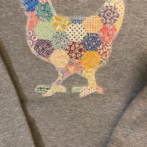 May include: A gray sweatshirt featuring a colorful patchwork chicken design. The chicken is composed of various fabric patterns, including floral, polka dot, and geometric shapes in shades of blue, pink, yellow, and green. The sweatshirt has a crew neck.