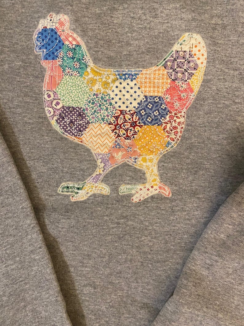 May include: A gray sweatshirt featuring a colorful patchwork chicken design. The chicken is composed of various fabric patterns, including floral, polka dot, and geometric shapes in shades of blue, pink, yellow, and green. The sweatshirt has a crew neck.