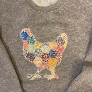 May include: A gray sweatshirt featuring a colorful patchwork chicken design. The chicken is made of various fabric patterns in shades of blue, yellow, red, and purple. The sweatshirt has a crew neck and long sleeves.