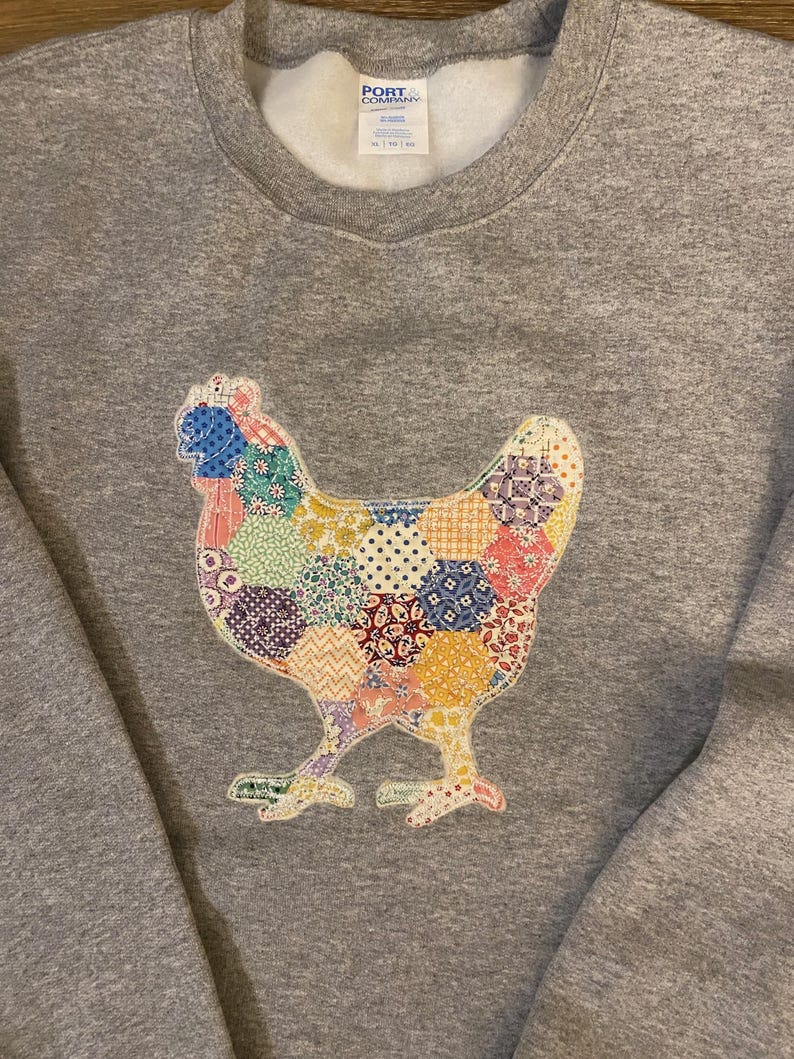 May include: A gray sweatshirt featuring a colorful patchwork chicken design. The chicken is made of various fabric patterns in shades of blue, yellow, red, and purple. The sweatshirt has a crew neck and long sleeves.