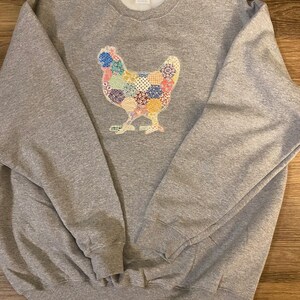 May include: A gray sweatshirt featuring a colorful patchwork chicken design. The chicken is made of various patterned fabrics in shades of blue, pink, yellow, and green. The sweatshirt has a crew neck and long sleeves.