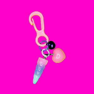 May include: A pink plastic keychain with a rainbow swirl charm, a pink heart charm with the letter "D" on it, and a purple bell charm.